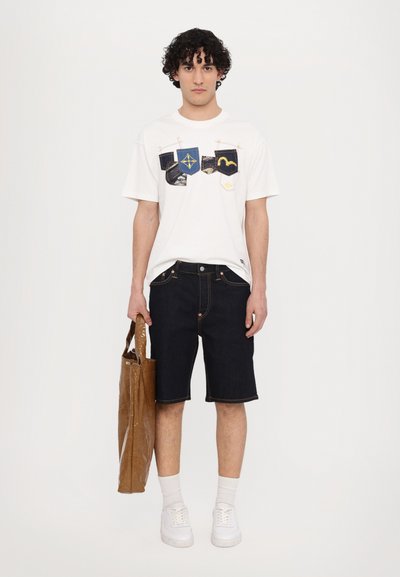 T-shirt blanc avec des motifs brodés en bleu et jaune. Short en denim foncé avec des coutures contrastées et un sac fourre-tout en simili cuir brun brillant. Baskets blanches.