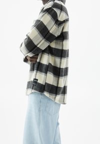 Persoon in een zwart-wit geruite jas met zijn armen gekruist, draagt lichtblauwe jeans tegen een witte achtergrond.