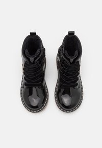 Botines tobilleros de charol negro con forro texturizado, punteras redondas y costuras blancas a contraste a lo largo de la suela. Presentan cordones y una cremallera lateral.