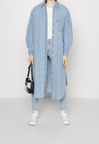 Vestido de camisa de mezclilla en azul claro, con botones en el frente, bolsillo en el pecho y mangas largas. Combinado con jeans claros y zapatillas blancas.