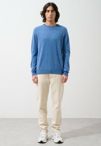 Blauer gestrickter Pullover mit rundem Ausschnitt, kombiniert mit hellen Chinos und weißen Sportschuhen. Einfaches Design und weiche Textur.