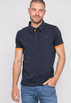 Jeune homme aux cheveux courts portant un polo bleu marine avec des bords des manches orange et un jean bleu clair, debout avec une main dans la poche.
