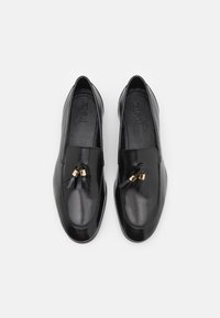 Paire de mocassins en cuir noir avec pompons et détails dorés vus de dessus sur fond blanc.