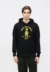 Čierna mikina s kapucňou s predným vreckom a žltou grafickou potlačou. Má šnúrky na stiahnutie a rebrované manžety, vyrobená z mäkkého bavlneného materiálu.