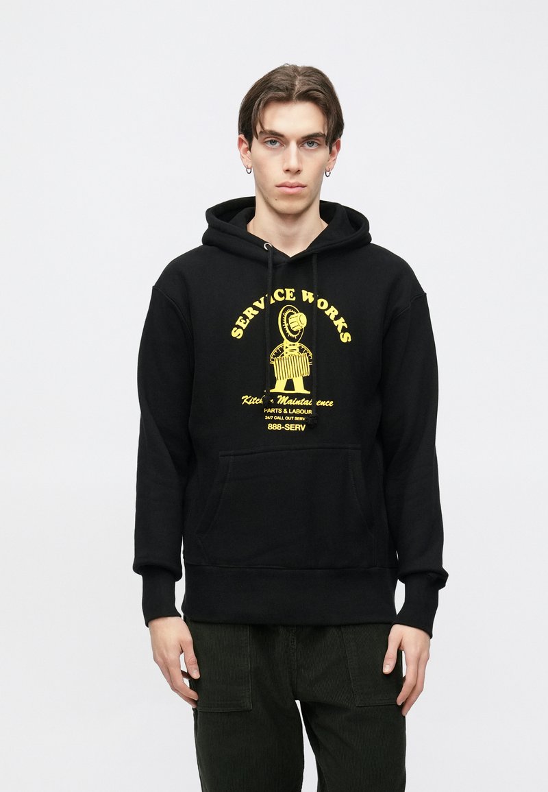 Čierna mikina s kapucňou s predným vreckom a žltou grafickou potlačou. Má šnúrky na stiahnutie a rebrované manžety, vyrobená z mäkkého bavlneného materiálu.