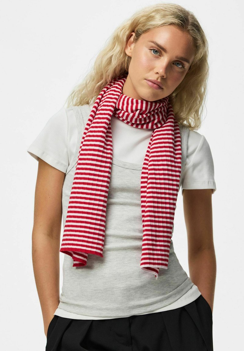 Jeune femme aux longs cheveux blonds portant un t-shirt blanc superposé d'un débardeur gris et une écharpe rayée rouge et blanche.