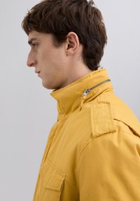 Ung mand i gul jakke med høj krave og lynlåsdetaljer, vist i sideprofil mod en ensfarvet lys baggrund.