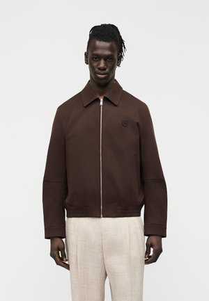 Mężczyzna ubrany w ciemnobrązową kurtkę z kołnierzem i jasnobeżowe, teksturowane spodnie, stojący na tle jednolitego jasnego tła.