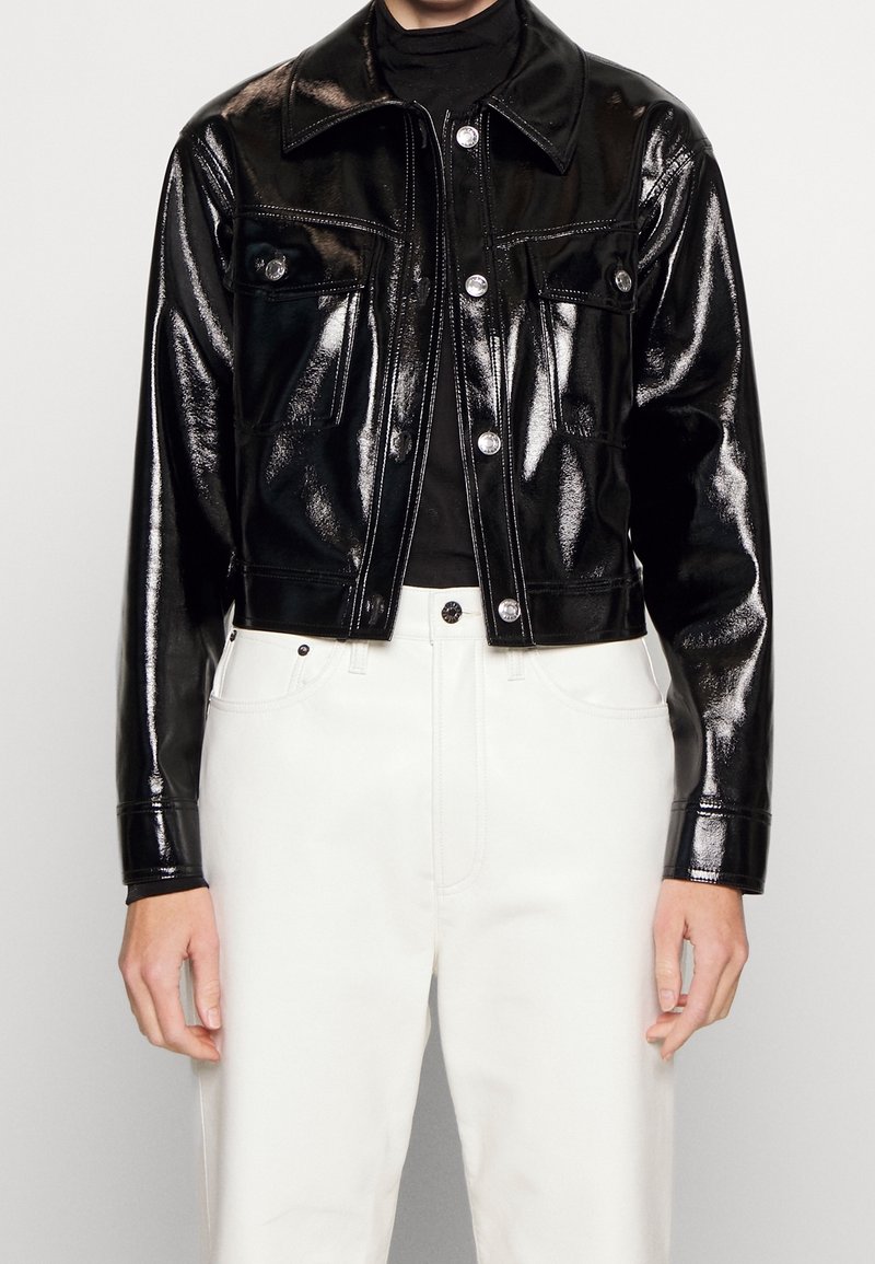 Veste noire brillante à boutons avec poches poitrine portée par-dessus un haut noir et un pantalon blanc, visible de la moitié du torse aux mains.