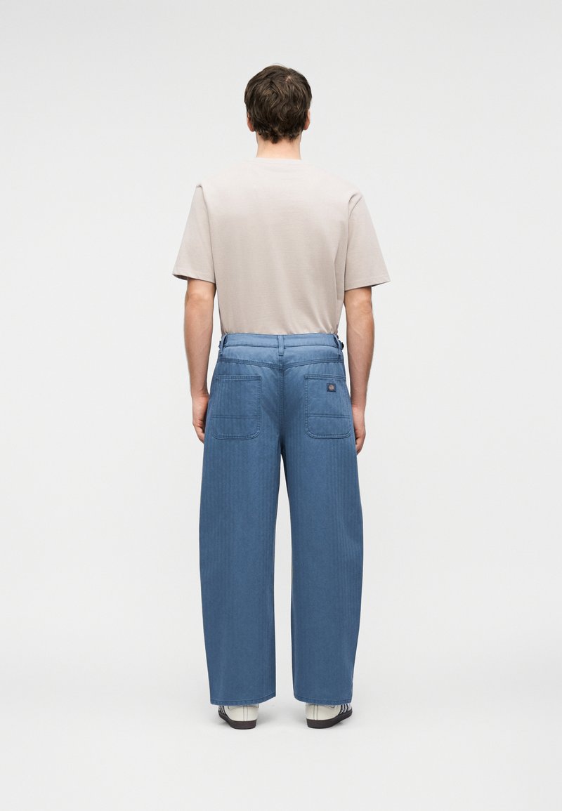 Man standing facing away, wearing a beige short-sleeve shirt, wide-leg blue pants with back pockets, and white sneakers with black stripes.