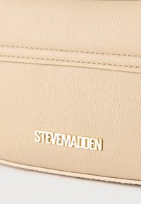 Επιφάνεια κρέμας με υφή δέρματος και χρυσό λογότυπο "Steve Madden" σε κοντινή λήψη τσάντας.