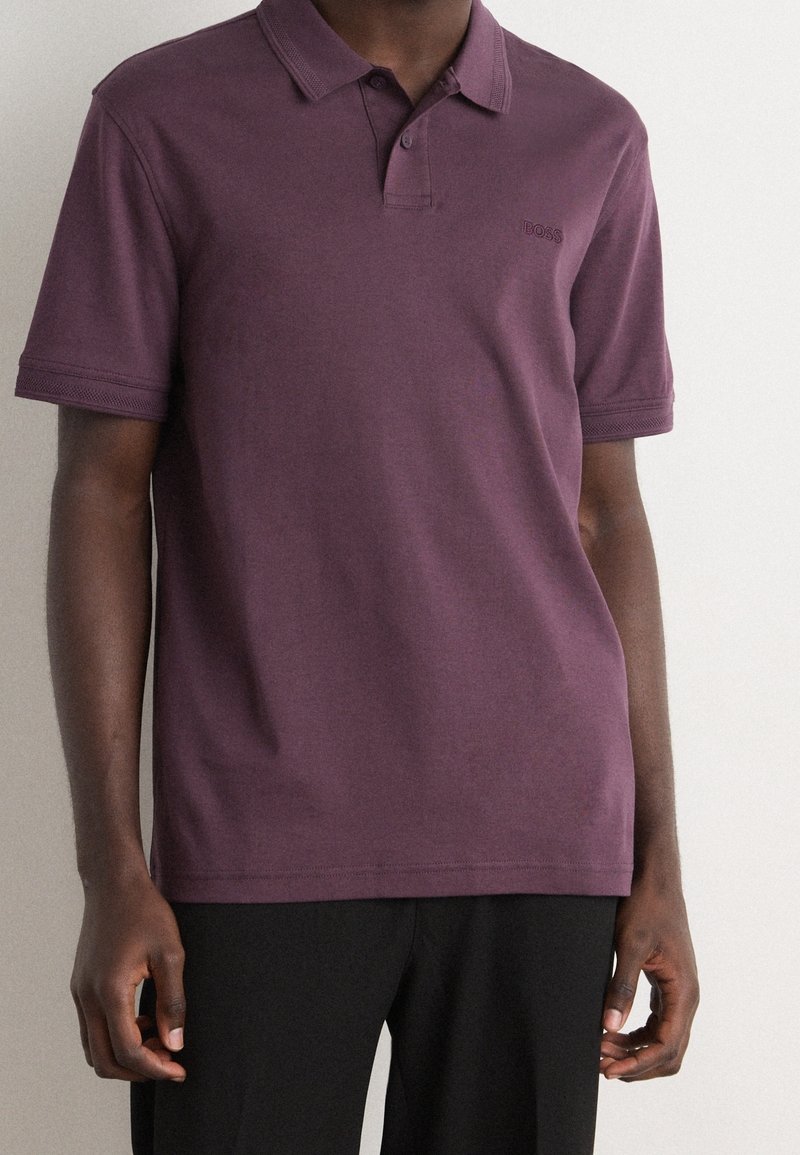 Homme portant un polo à manches courtes de couleur prune avec un logo discret sur la poitrine, associé à un pantalon noir sur fond clair.