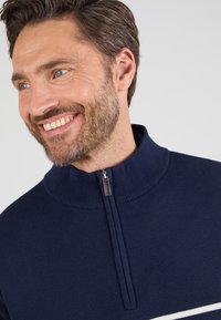 Pull en maille bleu marine avec col montant et zip quart. Présente une bande horizontale blanche sur la poitrine. Texture douce et design décontracté.
