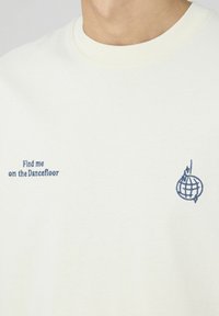 T-shirt en coton crème avec un col rond. Présente un texte brodé bleu : "Retrouve-moi sur la piste de danse" et un design de globe.