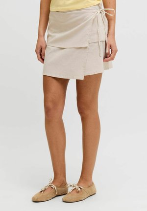 Mini-jupe portefeuille beige avec détail noué, portée avec des ballerines beiges sur une personne debout sur un fond uni.