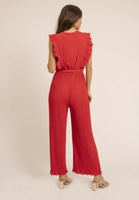 Combinaison rouge plissée avec des manches à volants et une taille cintrée. Jambes larges à coupe ample et ourlets festonnés. Portée avec des talons à lanières.