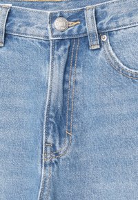 Gros plan sur un jean en denim bleu clair montrant la ceinture, le bouton, les passants de ceinture, la braguette à glissière et la poche avant droite avec des détails de couture visibles.