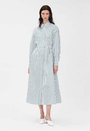 Femme portant une robe midi blanche avec des rayures verticales noires, aux longues manches, avec une ceinture nouée à la taille, se tenant debout avec des mules à talons blancs sur un fond blanc uni.