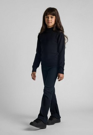 Niña joven con el cabello largo, vestida con un suéter de cuello alto negro, pantalones negros y zapatillas negras, de pie contra un fondo gris uniforme.