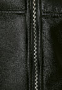 Närbild av svart strukturerat läder med en centrerad silverfärgad dragkedja i metall och sydda horisontella sömmar nära toppen.