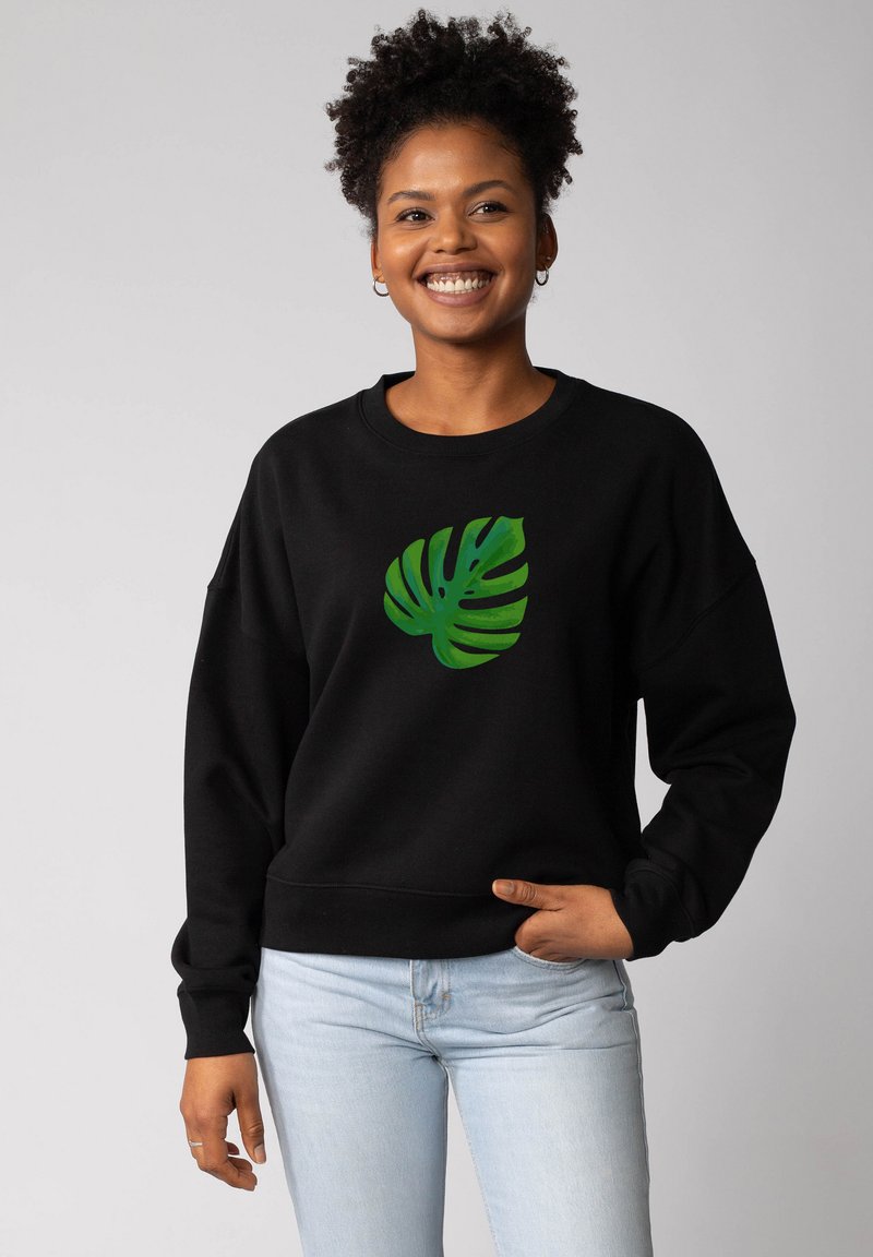 Sweatshirt noir avec un graphique de feuille de monstera verte au centre. Texture douce, coupe décontractée et poignets côtelés. Porté avec un jean bleu clair.