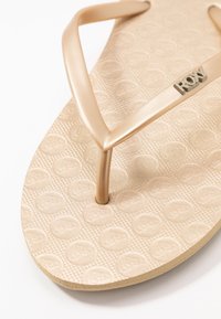 Beige flip-flop sandal with a textured footbed, featuring a glossy strap and embossed logo, highlighting a simple design and lightweight material.