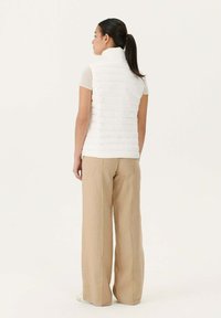 Chaleco acolchado blanco con cuello alto, vista trasera, llevado sobre una blusa de manga corta con canalé, combinado con pantalones de pierna ancha beige. Textura suave.
