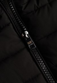 Svart puffjacka med dragkedjedetalj och silverfärgad metalldragare, med en texturerad svart dragkedja och "SUPERDRY"-logotyp på dragkappen.