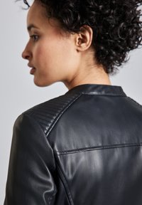 Veste en cuir noir avec une texture matelassée sur les épaules, finition lisse et design ajusté. Coutures visibles à l'arrière.