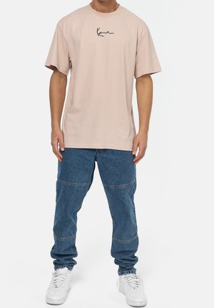 Vaaleanpunainen puuvillapaita, jossa on mustalla käsinkirjoitettu logo rinnassa, yhdistettynä sinisiin farkkuihin ja valkoisiin lenkkareihin.