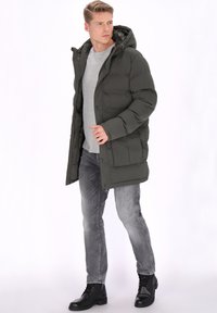 Ciemnozielona pikowana parka z kapturem, zapięciem na zamek i guziki, bocznymi kieszeniami oraz teksturowaną tkaniną. Noszona na szarej koszuli i dżinsach.