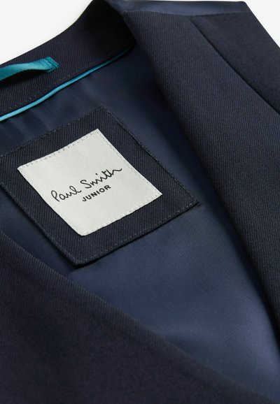 Mörkblå blazer tyg med en jämn textur; har en vit etikett med "Paul Smith JUNIOR" i svart kursiv stil. Utsmyckad med en turkos kant.