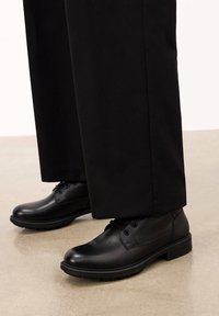 Mustad nahast kingad sileda tekstuuriga, ümmarguse toe ja paeltega disainiga. Paigutatud mustade pükste kõrvale heledale põrandale.