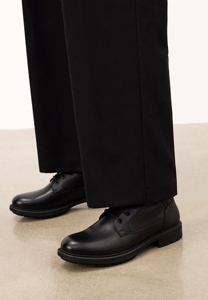 Mustad nahast kingad sileda tekstuuriga, ümmarguse toe ja paeltega disainiga. Paigutatud mustade pükste kõrvale heledale põrandale.