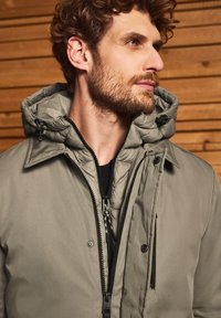 Ljusgrå isolerad jacka med avtagbar huva, dragkedjestängning och fickor med dragkedja. Slät textur med synliga sömdetaljer.