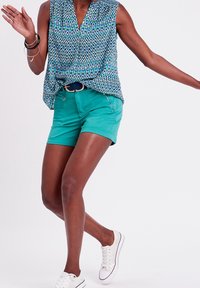 Farverig mønstrede ærmeløse top med V-udskæring, parret med teal shorts og hvide sneakers. Fashionable accessories inkluderer flere armbånd.
