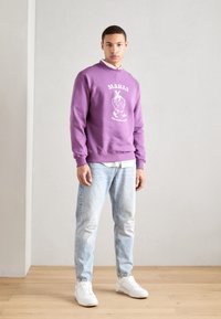 Lilla crewneck sweatshirt med hvit grafisk design, kombinert med lyseblå slitte jeans og hvite sneakers. Bakgrunn med tregulv.