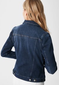 Tmavě modrá džínová bunda se rovného střihu, viditelným prošíváním, knoflíkovými detaily na manžetách a klasickým límcem. Zobrazen zadní pohled.