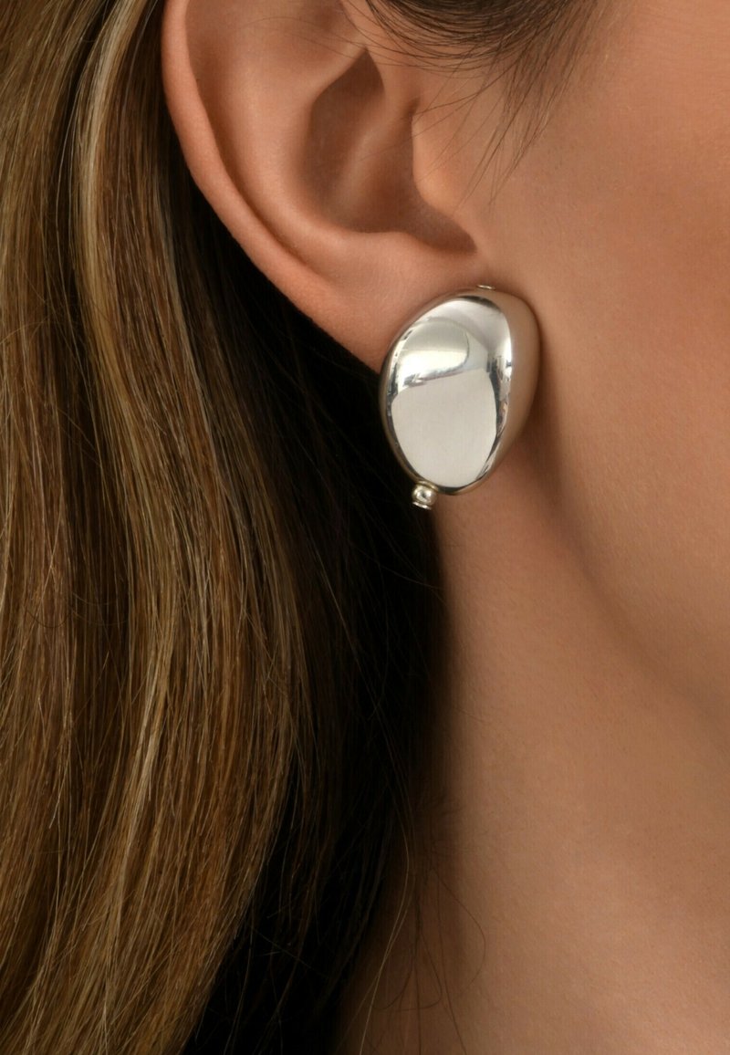 Oreille d'une femme avec un clou d'oreille ovale en argent lisse et brillant et des cheveux châtain clair visibles sur le côté.