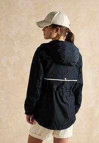 Jūras spēku ūdensnecaurlaidīga jakas ar pieguļošu jostu, kapuci un baltu akcentu lentu. Adīta cepure un gaiši šorti papildina apģērbu.