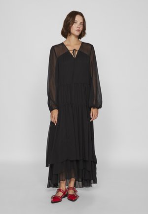 Black long-sleeve dress with a V-neck and tie detail, featuring a layered skirt design in sheer fabric. Paired with red flat shoes with studs.