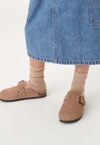 Brūnas zamšādas kurpes ar sprādzes detaļu, valkātas ar ribotiem bēša krāsas zeķēm, kopā ar džinsu svārkiem. Uz gaišas teksturētas virsmas.