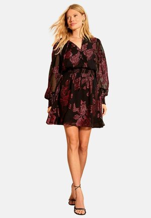 Svart klänning med blommigt mönster i burgundy-toner, långa genomskinliga ärmar, åtsittande midja och en utställd kjol. Bärs med svarta sandaler med remmar.