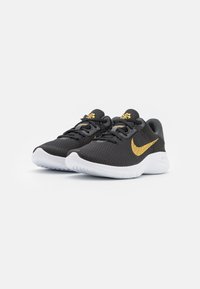 Une paire de chaussures de sport noires avec des semelles blanches, un logo Nike en or et un logo doré sur la languette, placées côte à côte sur un fond blanc.