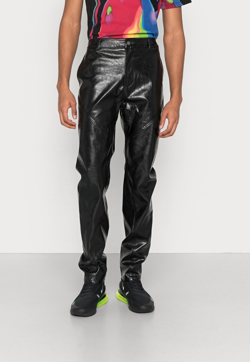 Musta tekonahkaverkkohousut suoralla leikkauksella, joissa on etutaskut ja kiiltävä pinta, yhdistettynä mustiin tossuihin ja värikkääseen yläosaan.