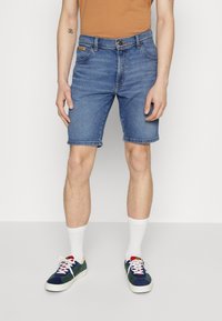 Shorts de mezclilla en azul claro con cortes rectos, cinco bolsillos y cierre de botón metálico. Combinados con calcetines blancos y zapatillas multicolores.