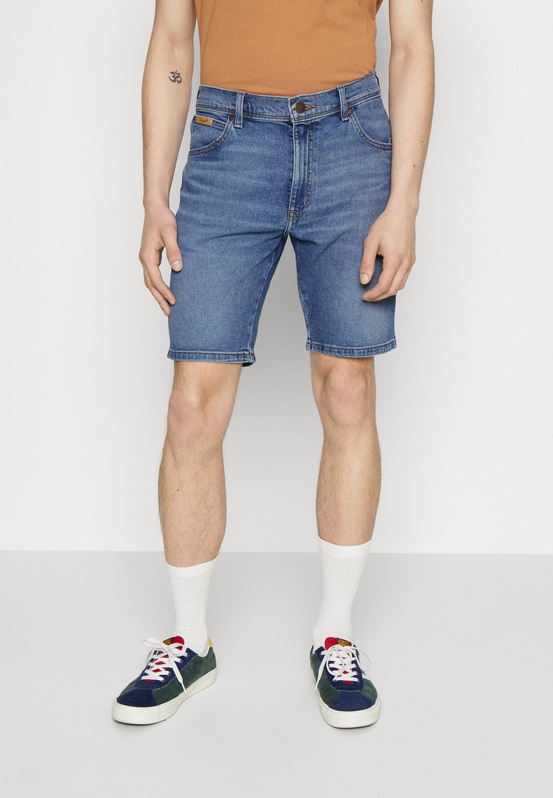 Shorts de mezclilla en azul claro con cortes rectos, cinco bolsillos y cierre de botón metálico. Combinados con calcetines blancos y zapatillas multicolores.