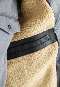 Capa exterior gris con un forro interior de felpa beige y detalles en cuero negro. Presenta una textura suave y secciones de materiales contrastantes.