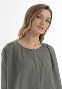 Blusa verde chiaro, con bottoni, dal colletto rotondo e maniche lunghe. Realizzata in tessuto morbido e testurizzato, con piccoli bottoni e dettagli arricciati.
