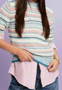 Personne portant un pull en tricot rayé sur une chemise à boutons rose et blanche à rayures, associé à un jean bleu, sur un fond violet.